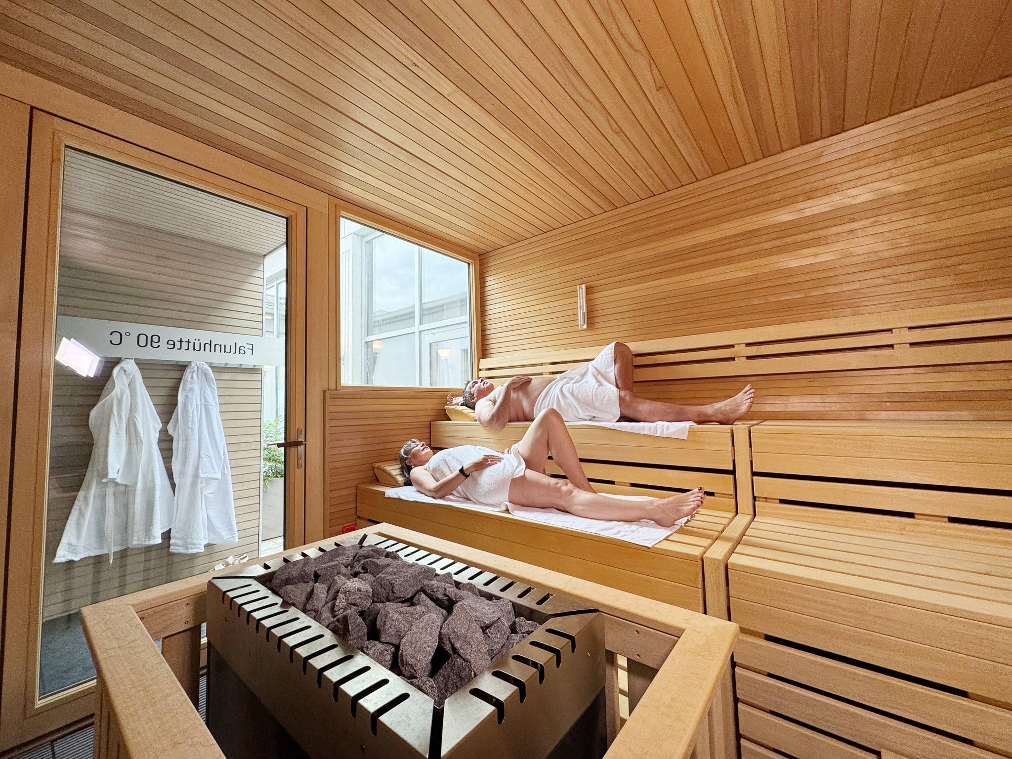 People relaxing in a wooden sauna, lying on two-tier benches with towels in a steam room salt-gray rocks on a heater in the foreground.