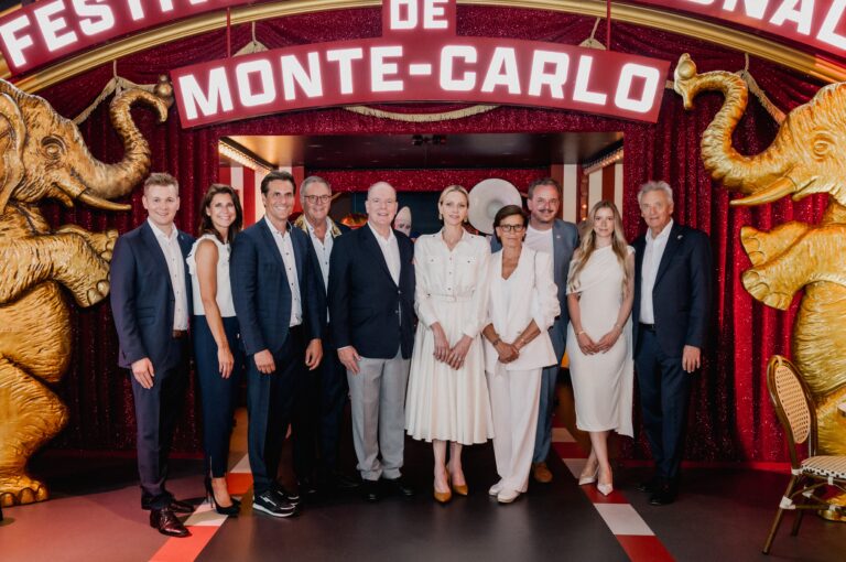 Group of professionals in white and navy posing at a Monte‑Carlo themed carnival entrance with gold elephant statues on each side.