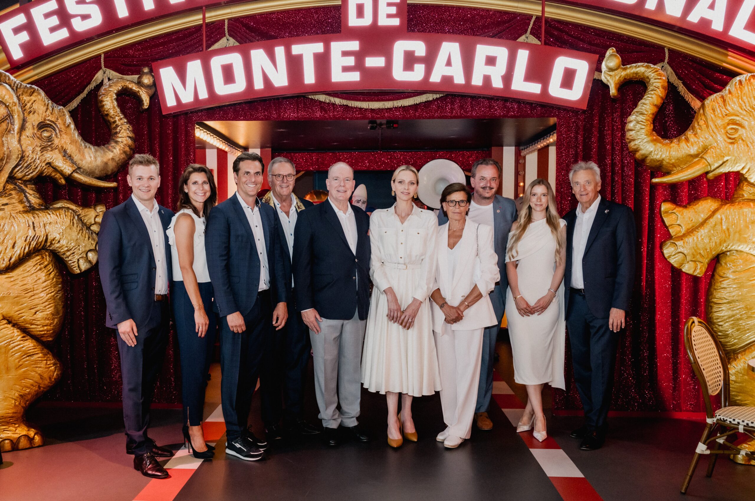 Group of professionals in white and navy posing at a Monte‑Carlo themed carnival entrance with gold elephant statues on each side.