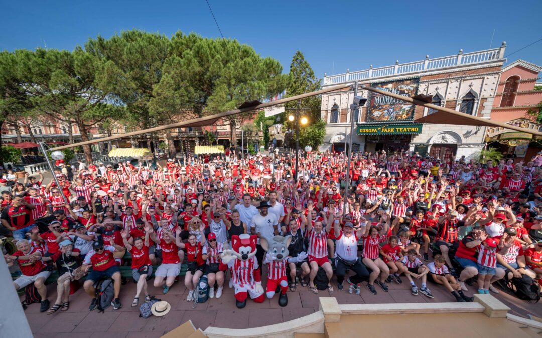 Massive crowd of fans in red-and-white striped shirts fills an outdoor plaza, many raising hands, with a theatre facade and trees in the background.