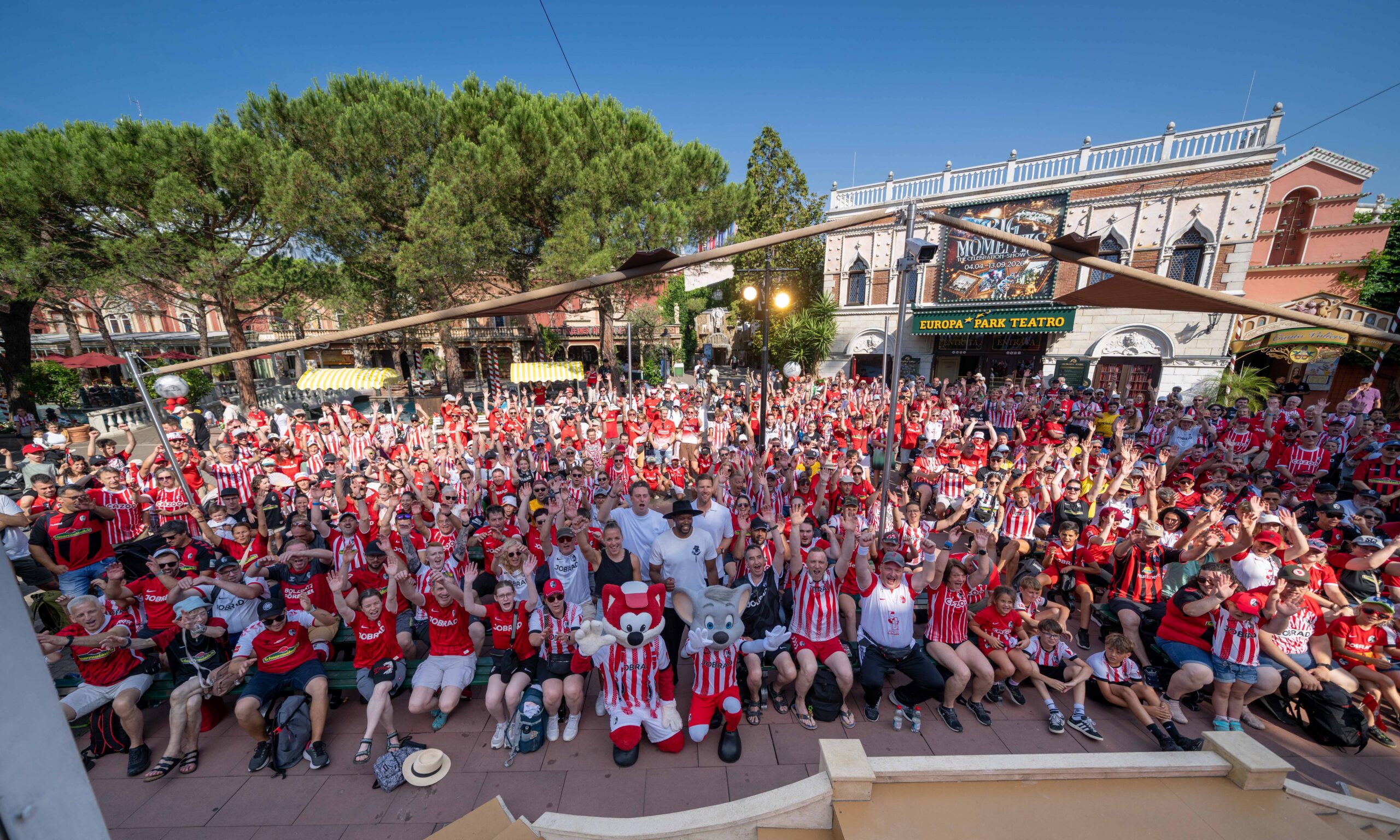 Massive crowd of fans in red-and-white striped shirts fills an outdoor plaza, many raising hands, with a theatre facade and trees in the background.