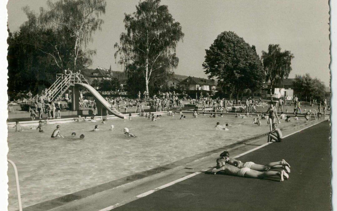 Crowded outdoor public pool with swimmers, sunbathers, a curved slide, and a diving area; trees and houses in the background.