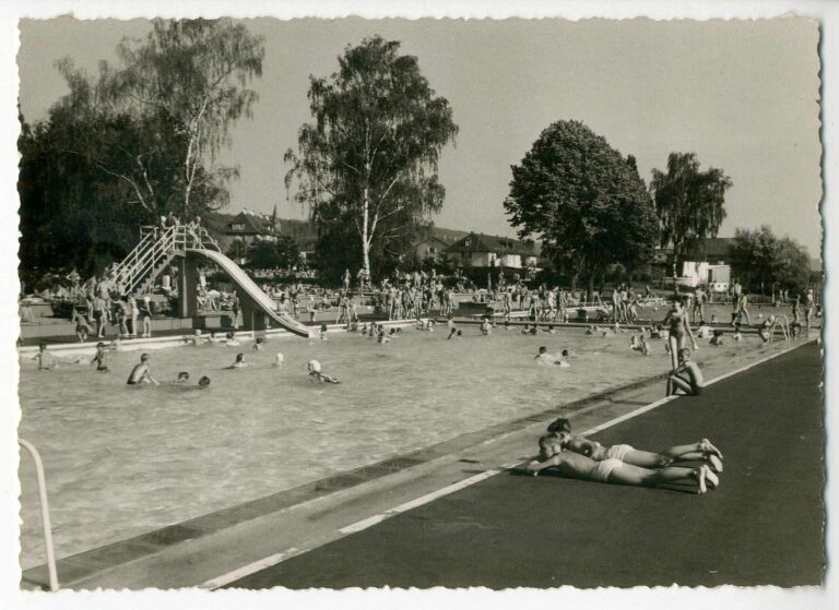 Crowded outdoor public pool with swimmers, sunbathers, a curved slide, and a diving area; trees and houses in the background.