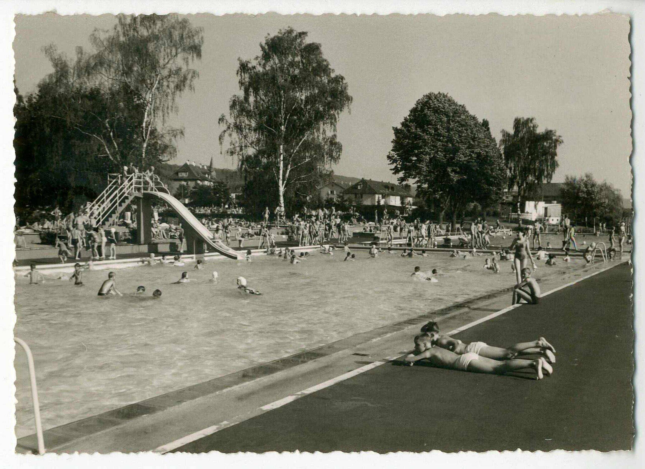 Crowded outdoor public pool with swimmers, sunbathers, a curved slide, and a diving area; trees and houses in the background.