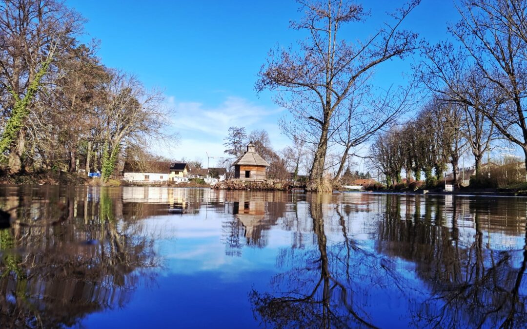 Calm river reflecting bare trees and a small wooden cottage with a shingled roof under a clear blue sky.