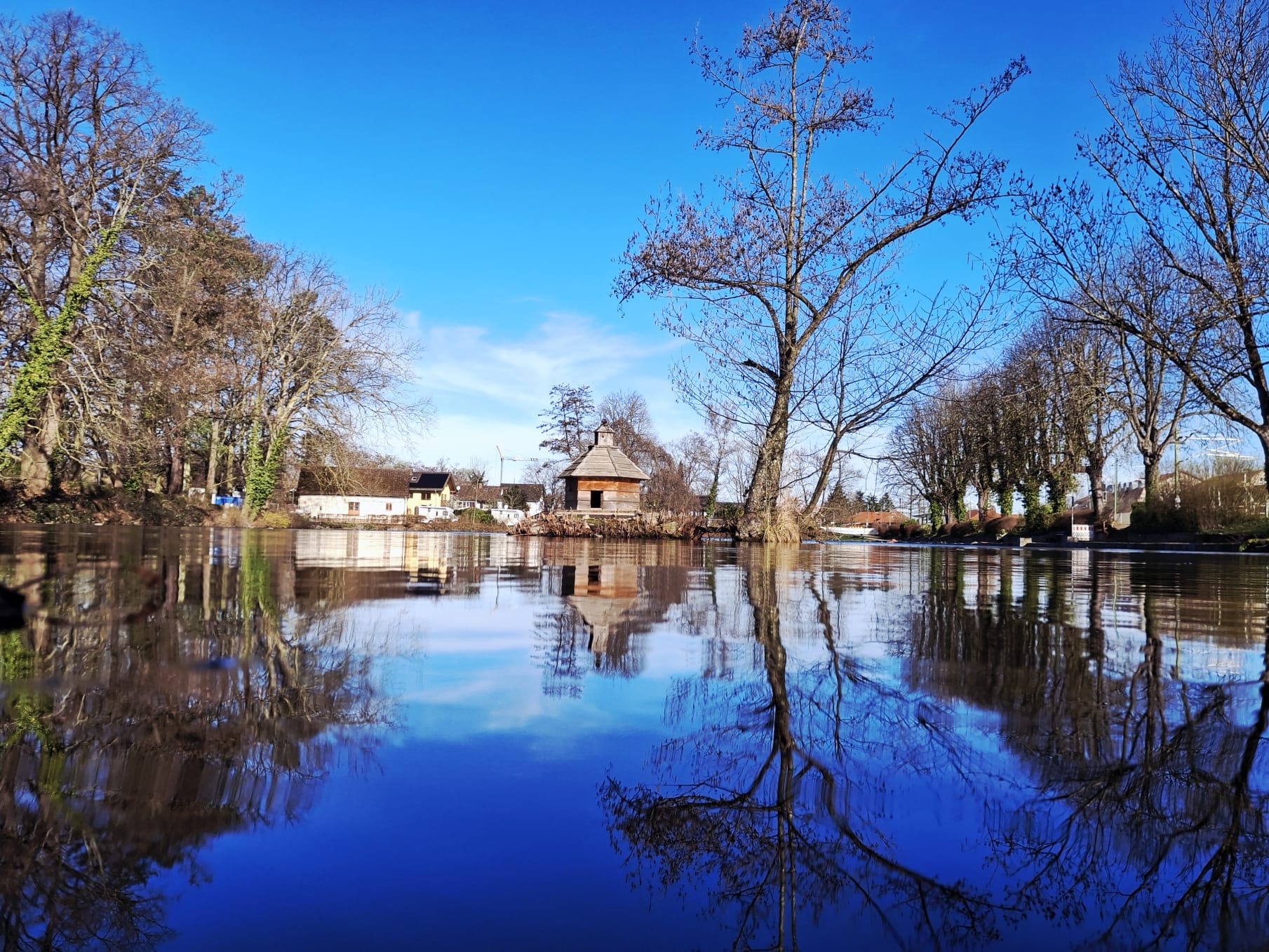 Calm river reflecting bare trees and a small wooden cottage with a shingled roof under a clear blue sky.
