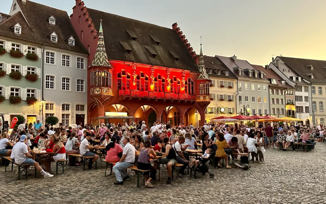 Crowded outdoor beer garden in a cobblestone square with colorful historic buildings and a bright red illuminated facade.",