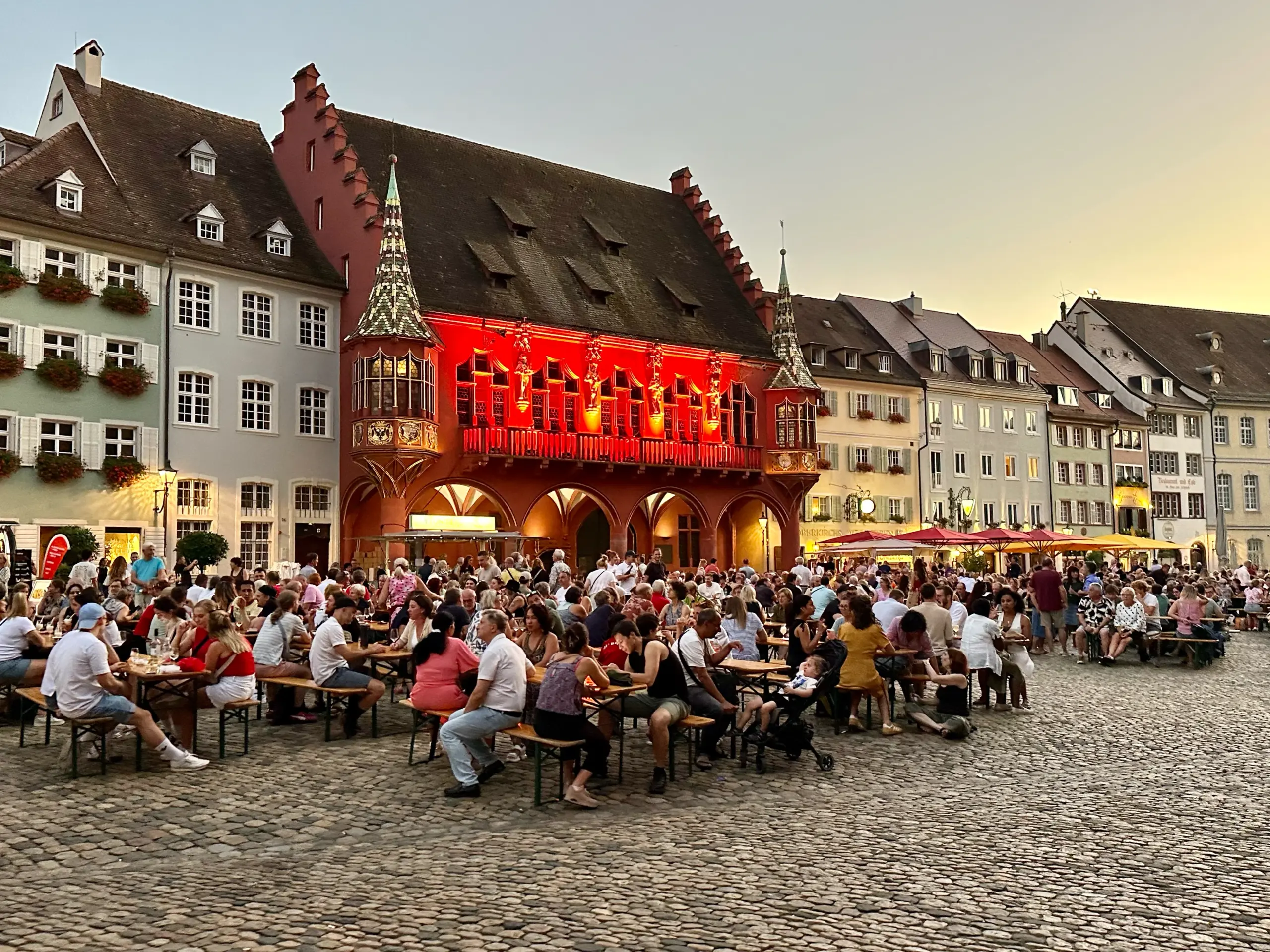 Crowded outdoor beer garden in a cobblestone square with colorful historic buildings and a bright red illuminated facade.",
