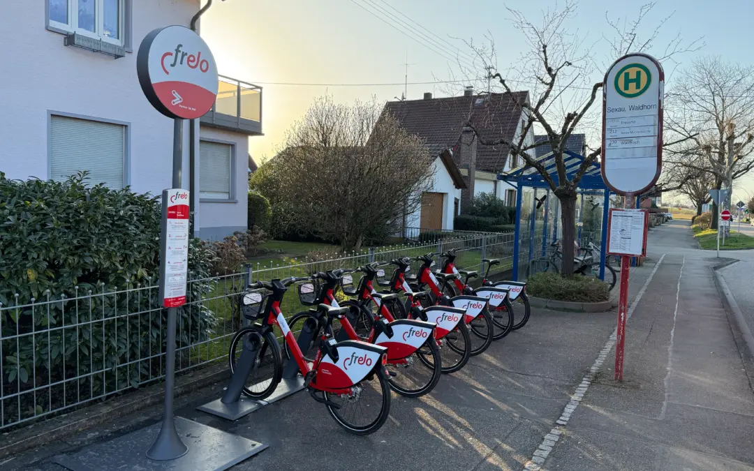 Row of red and white Frelo rental bicycles docked at a bike-sharing station along a sidewalk, with a bus stop sign in the background.