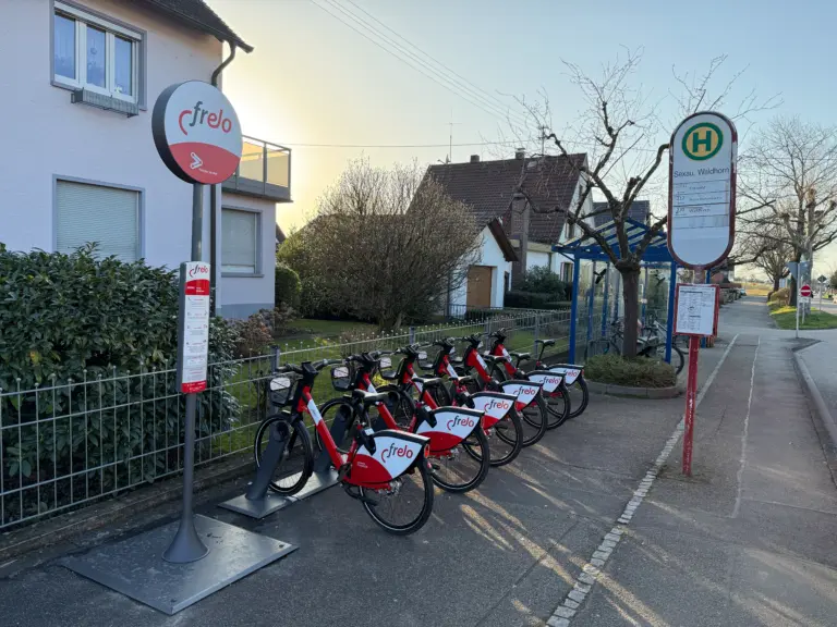 Row of red and white Frelo rental bicycles docked at a bike-sharing station along a sidewalk, with a bus stop sign in the background.