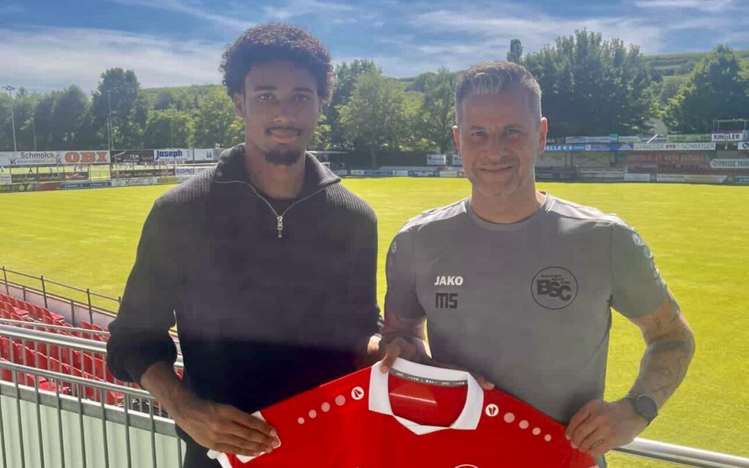 Two men standing on a stadium terrace, posing with a red football jersey that reads 'Fürstenberg' and shows a team logo. sunny day with green field behind them.
