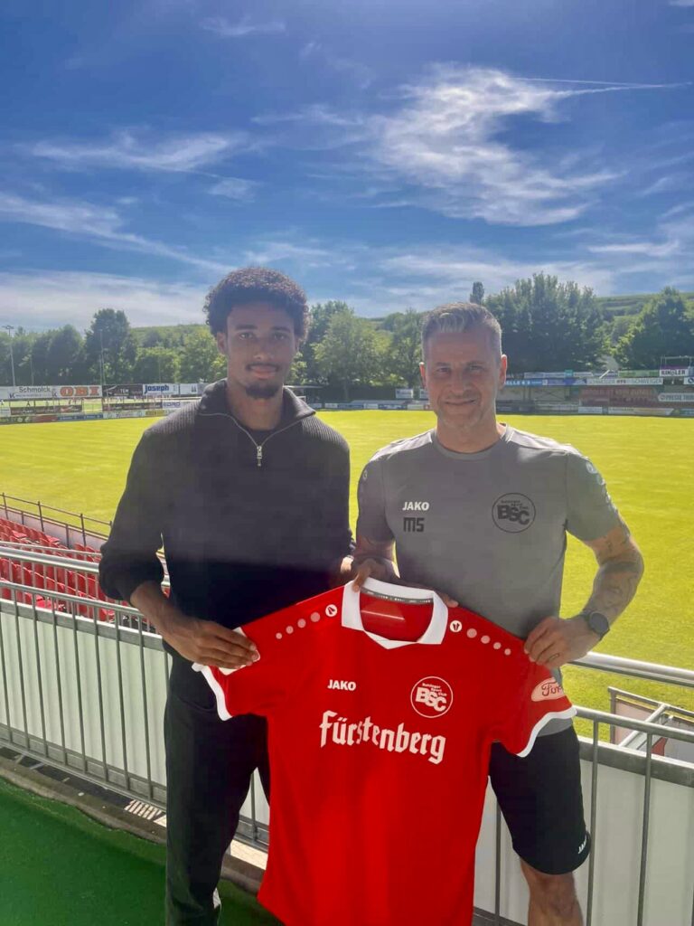 Two men standing on a stadium terrace, posing with a red football jersey that reads 'Fürstenberg' and shows a team logo. sunny day with green field behind them.