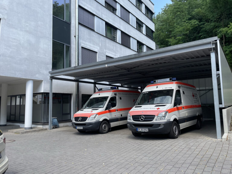 Two white ambulances with orange stripes parked under a metal carport in front of a gray building with large windows.