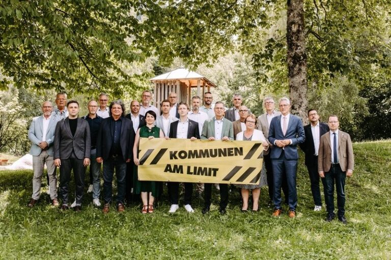 Group of adults in business attire posing outdoors, holding a yellow banner reading 'KOMMUNEN AM LIMIT' in a park.