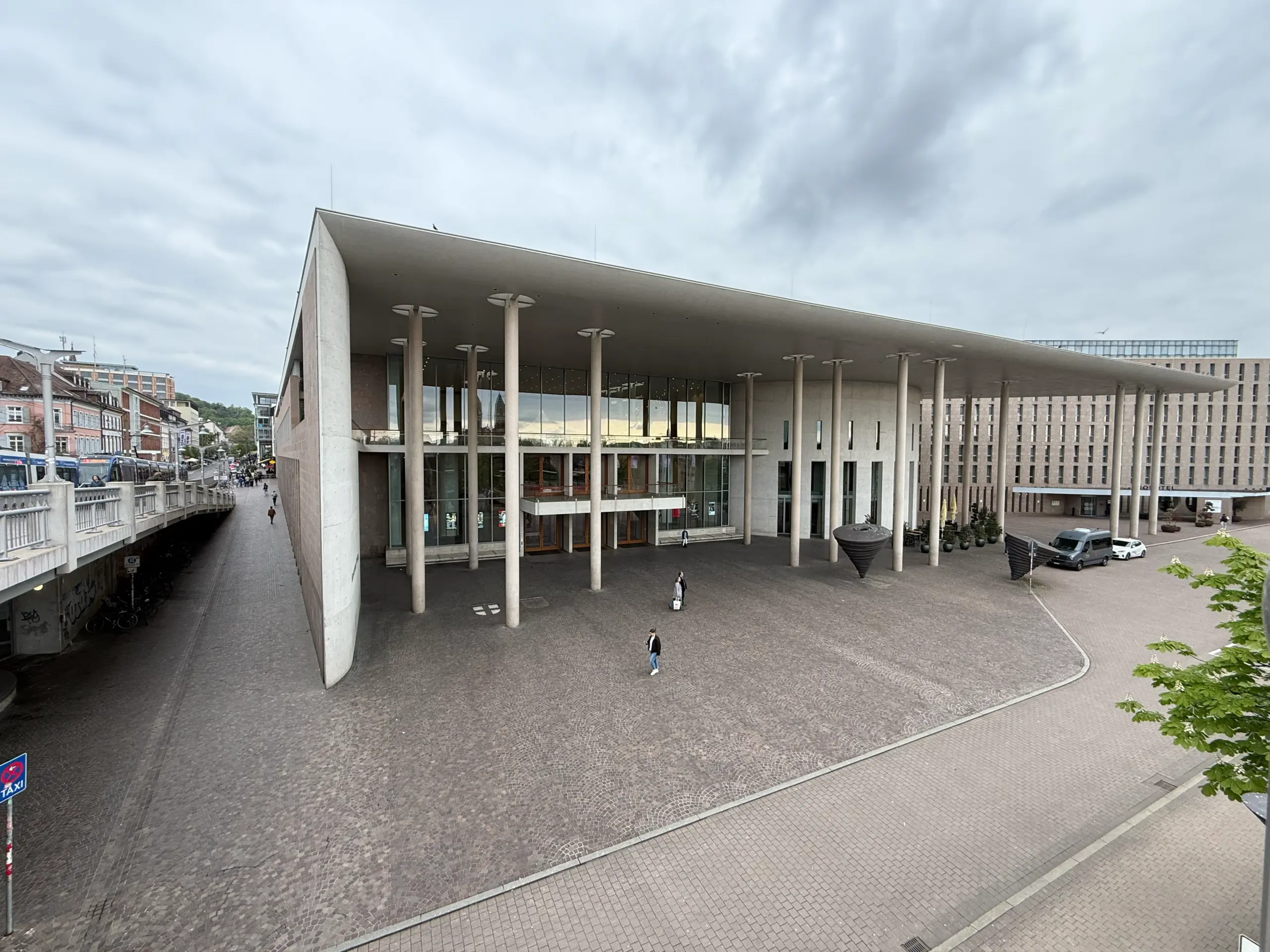 Modern public building with a long flat roof supported by white columns along a cobblestone plaza, under a cloudy sky