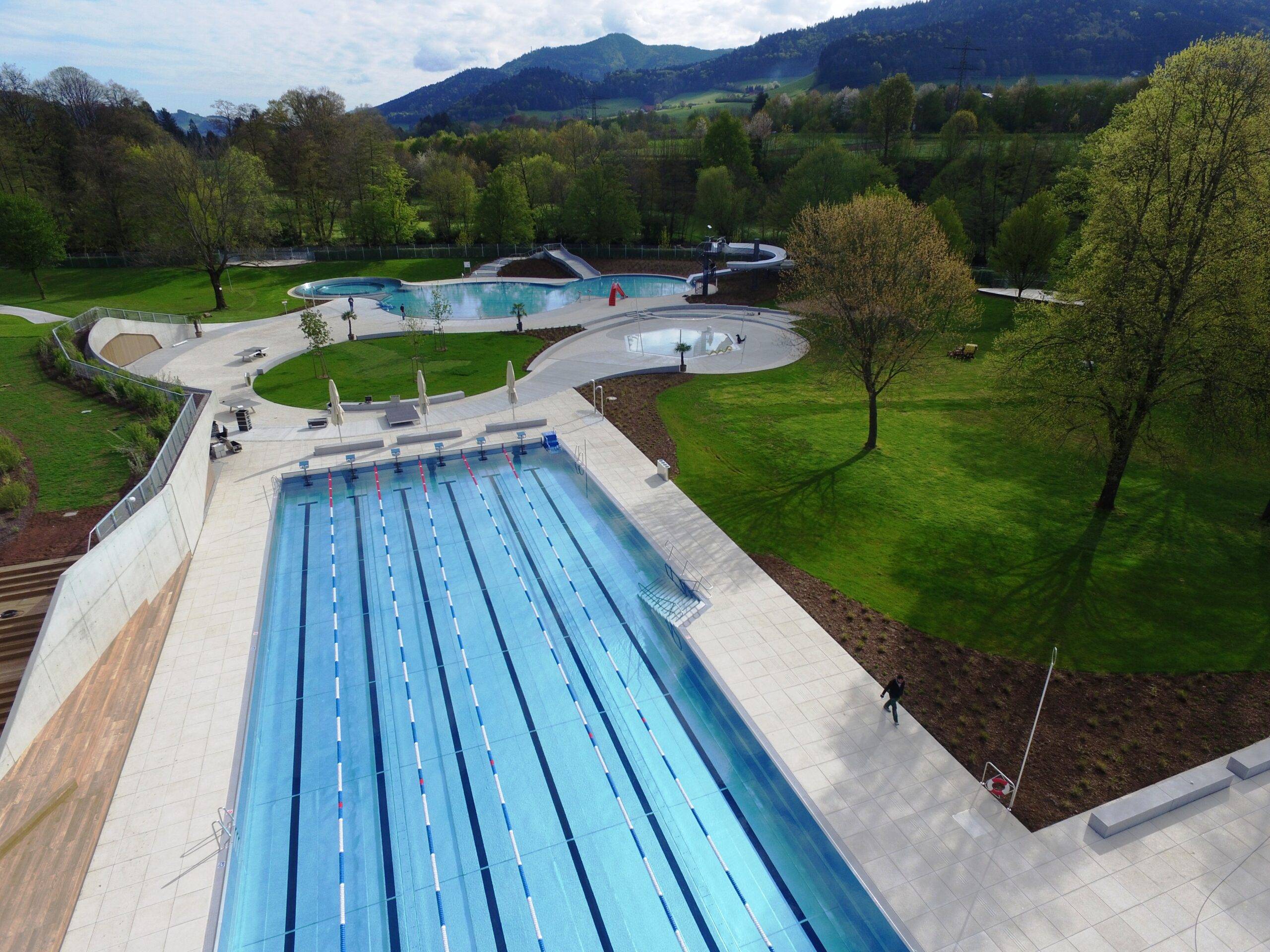 Aerial view of a rectangular outdoor swimming pool complex with multiple lanes, adjacent sunbathing area, and green hills in the background.