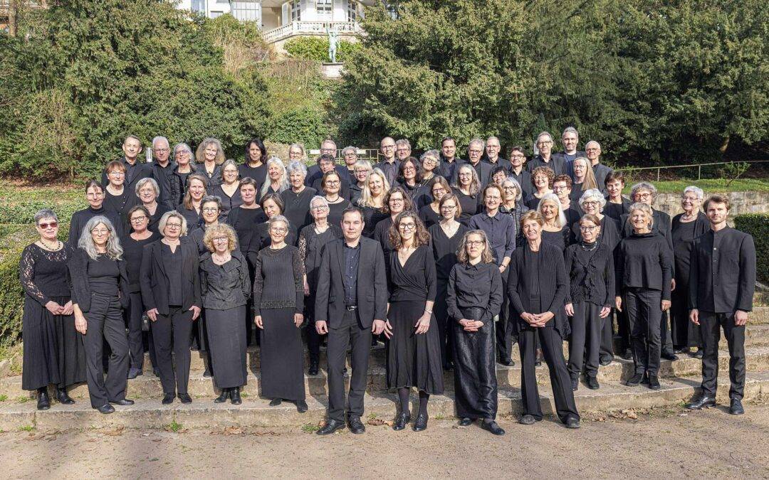 Large choir group dressed in black posing on stone steps outdoors with greenery behind them.