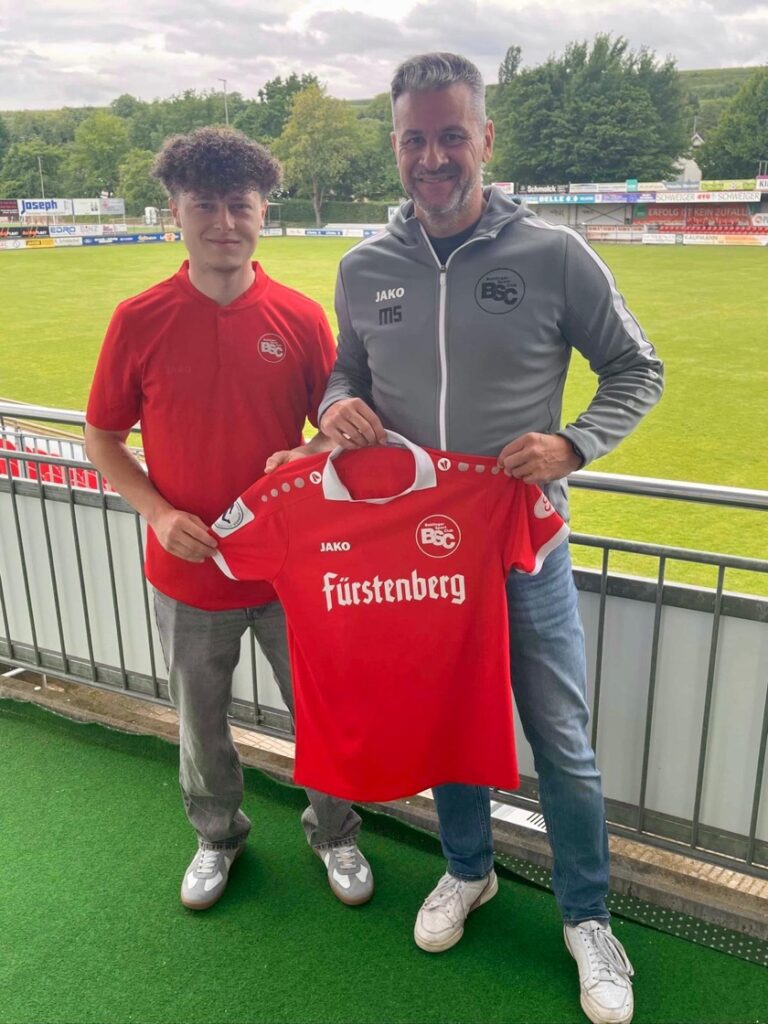 Two men stand on a stadium terrace holding up a red football jersey with 'Fürstenberg' sponsor, smiling at the camera.