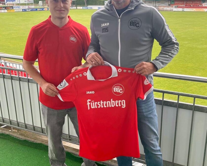 Two men stand on a stadium terrace holding up a red football jersey with 'Fürstenberg' sponsor, smiling at the camera.