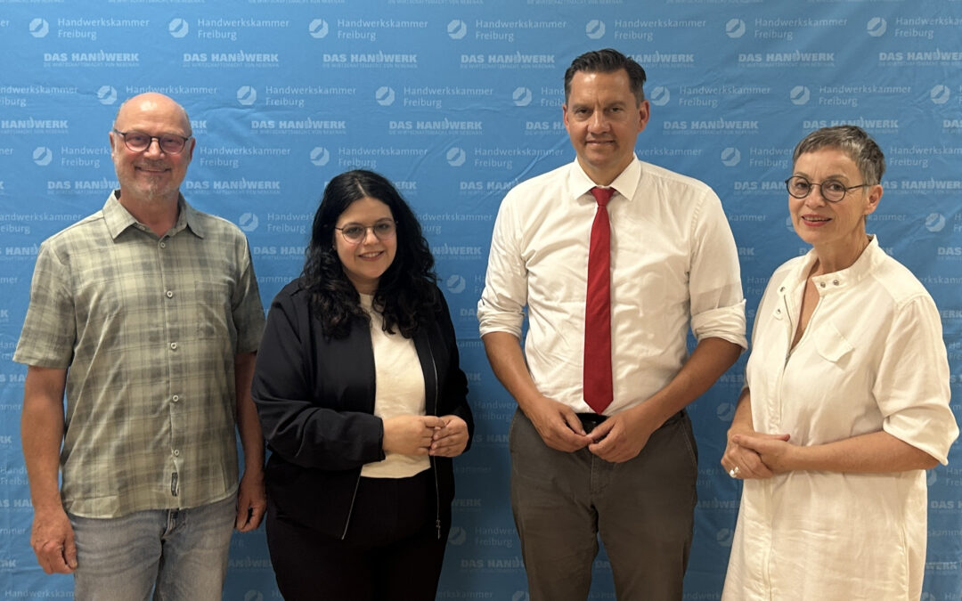 Group of four adults posing together in front of a blue backdrop with white 'DAS HANDWERK Freiburg' logos.