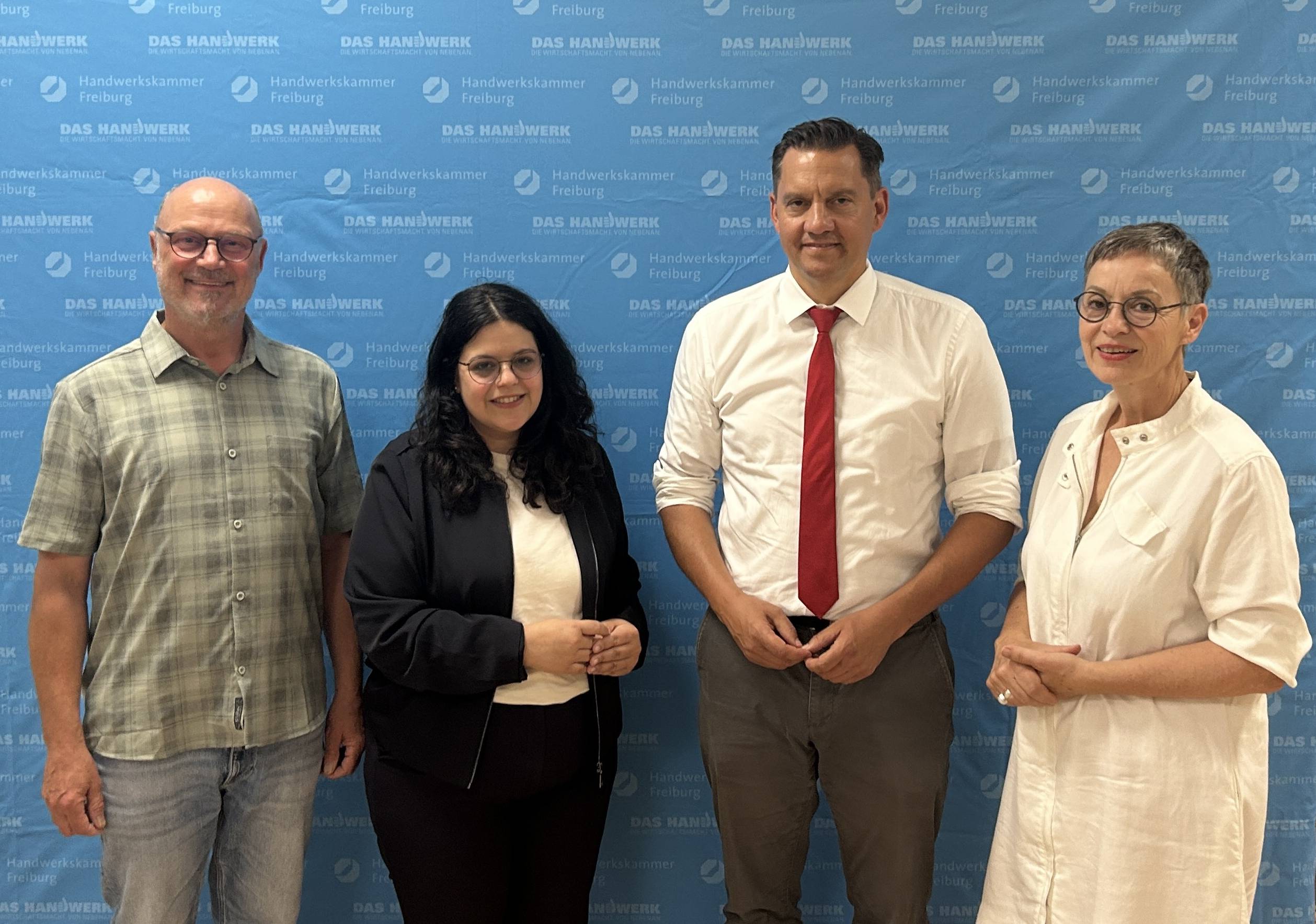 Group of four adults posing together in front of a blue backdrop with white 'DAS HANDWERK Freiburg' logos.