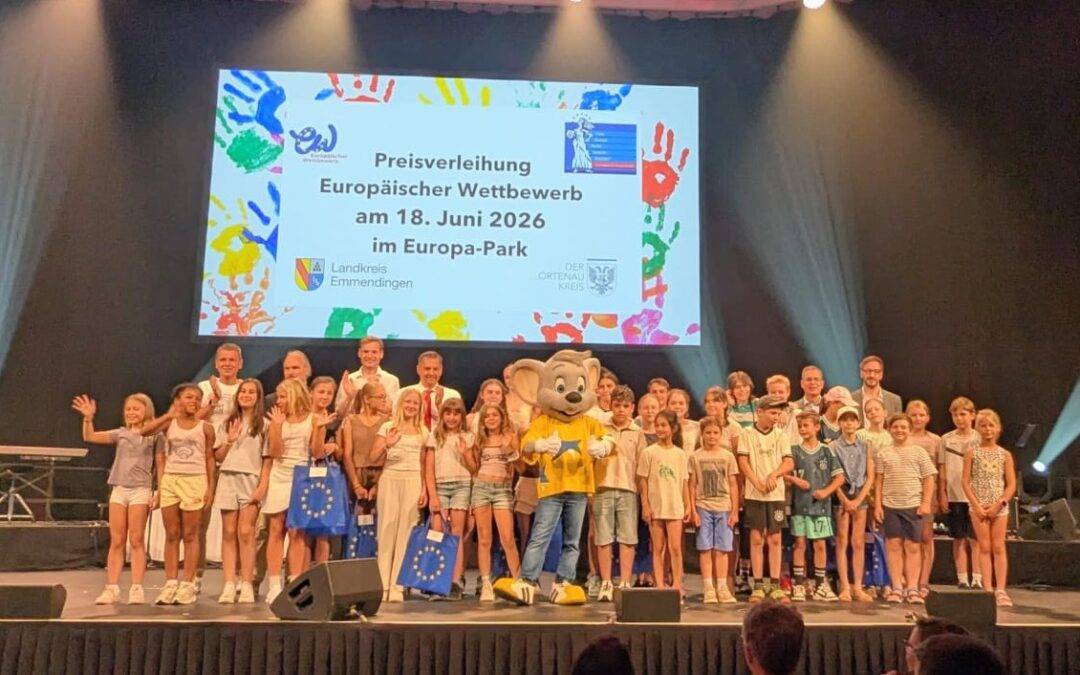 Children and adults posing on a theater stage with a yellow mascot; a large screen behind shows the German award event title, suggesting an award ceremony .