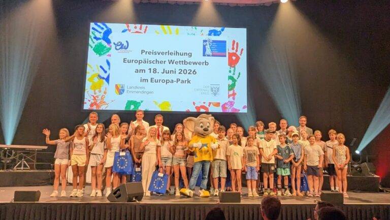 Children and adults posing on a theater stage with a yellow mascot; a large screen behind shows the German award event title, suggesting an award ceremony .