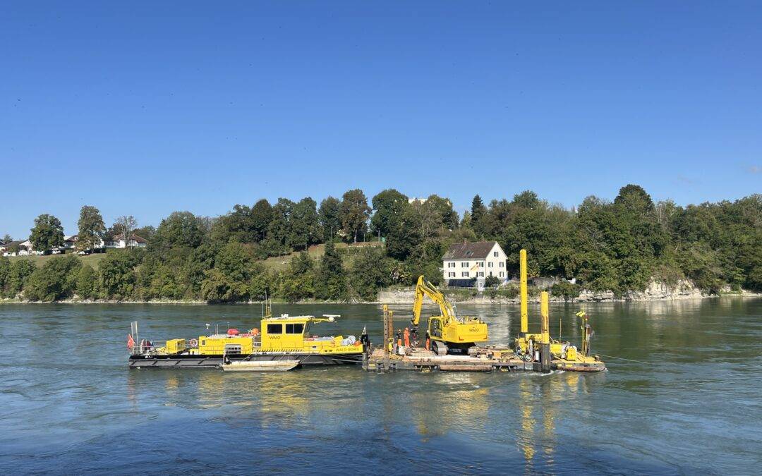 Yellow construction barge and machinery on a calm river with a tree-lined shore and houses in the background.