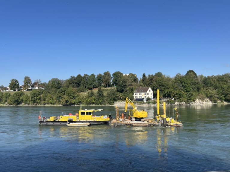 Yellow construction barge and machinery on a calm river with a tree-lined shore and houses in the background.