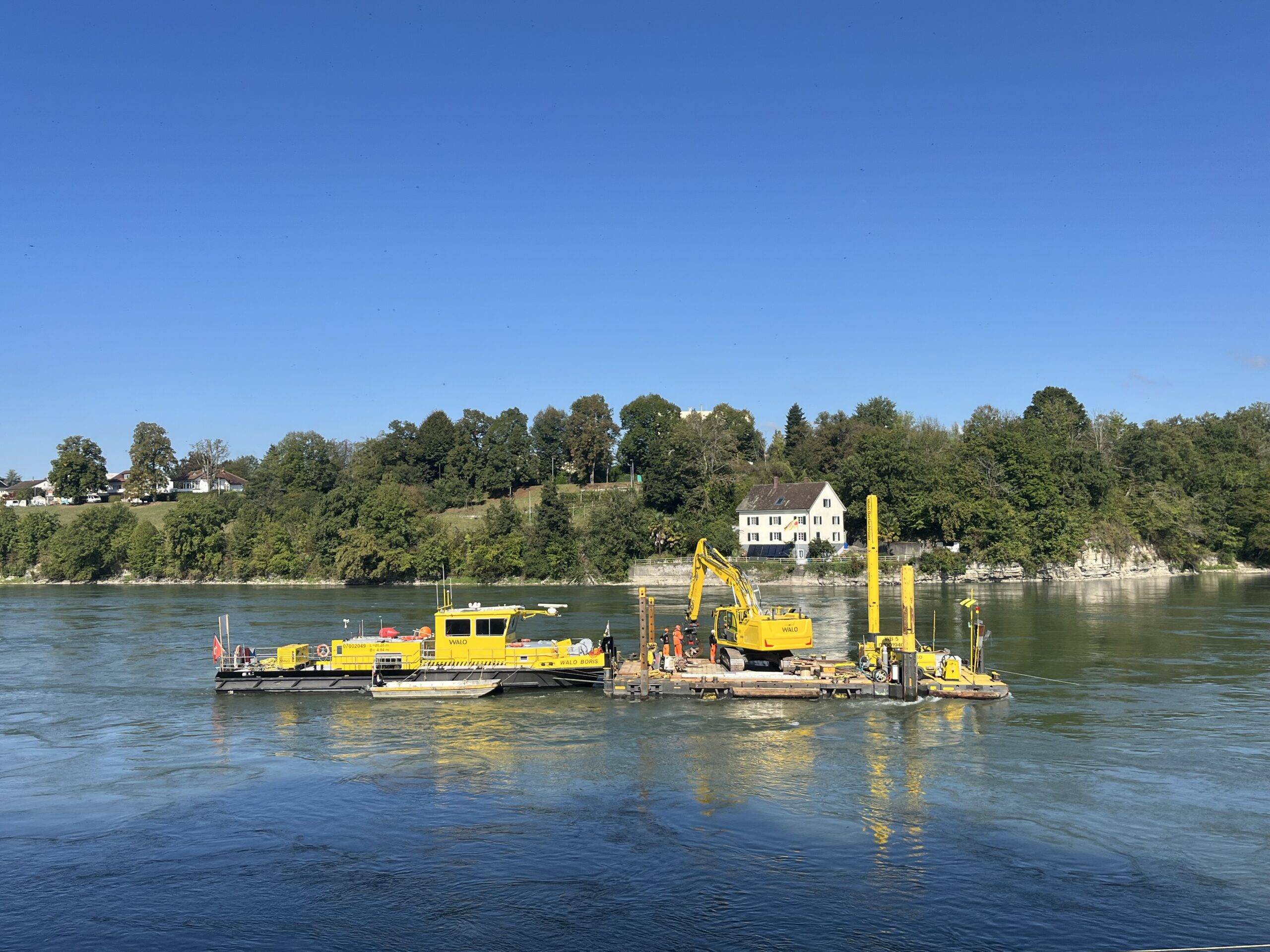 Yellow construction barge and machinery on a calm river with a tree-lined shore and houses in the background.
