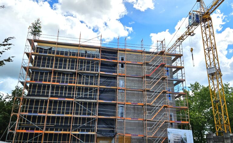 Multi-story building under construction with orange scaffolding and a tall yellow crane against a bright blue sky.