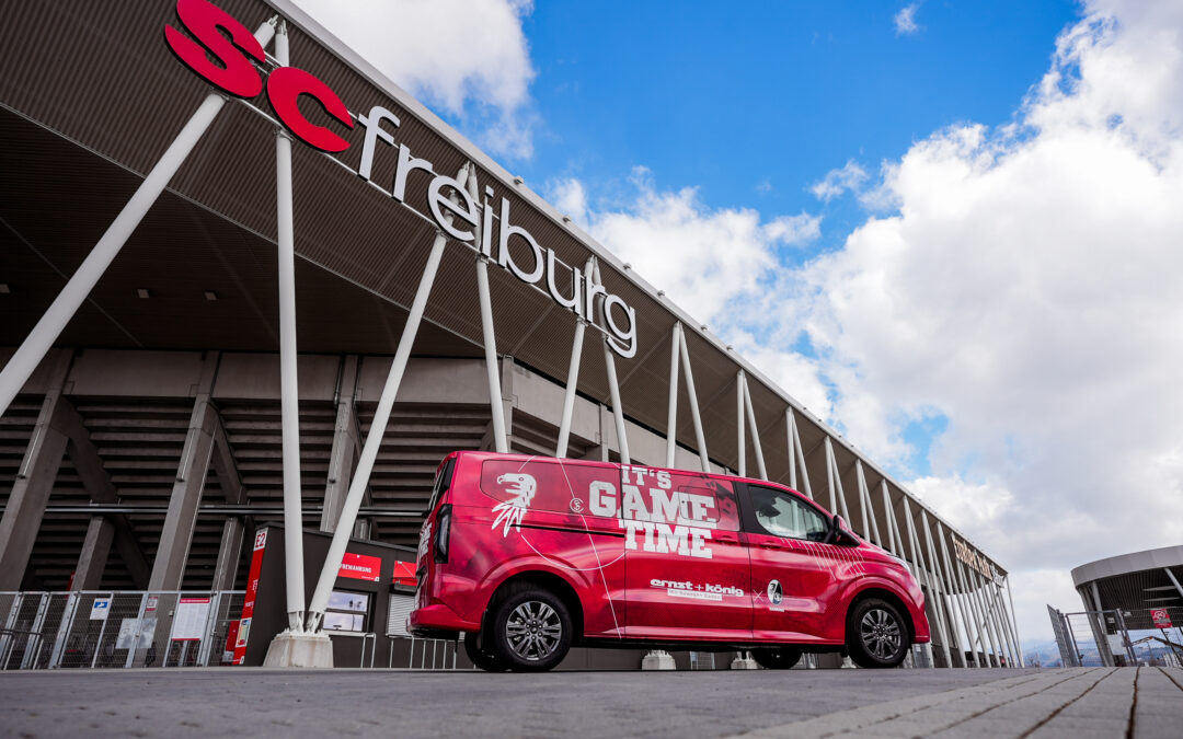 Red promotional van with 'It's GAME TIME' slogan parked outside a modern stadium under a blue sky.