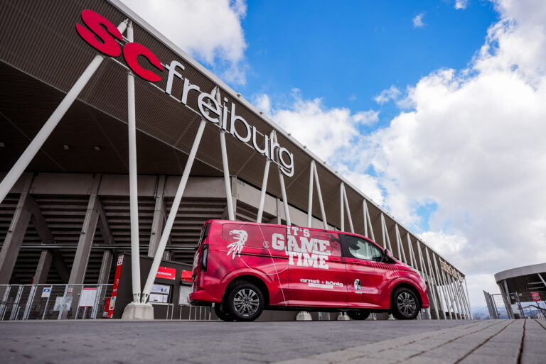 Red promotional van with 'It's GAME TIME' slogan parked outside a modern stadium under a blue sky.
