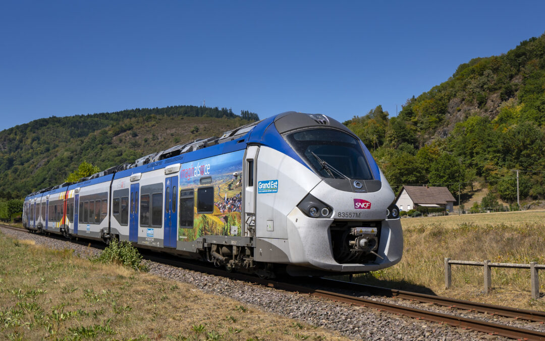 Modern blue and silver passenger train on tracks in a sunny rural landscape with forested hills and a clear blue sky behind it.