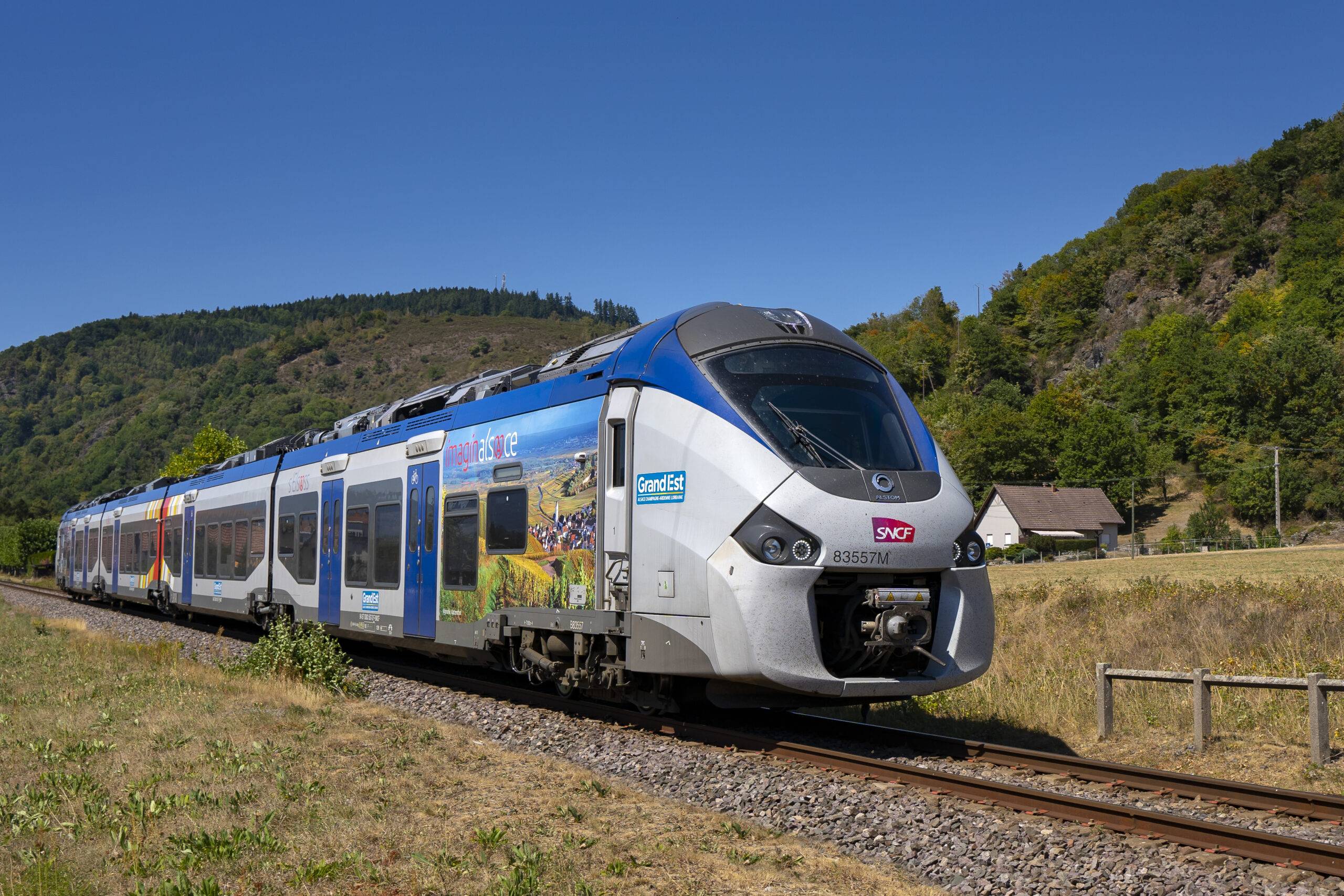 Modern blue and silver passenger train on tracks in a sunny rural landscape with forested hills and a clear blue sky behind it.