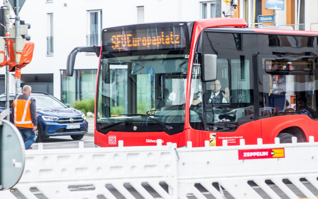 Red city bus with DB logo front and center, driver visible through the window, urban street scene with construction barriers nearby.