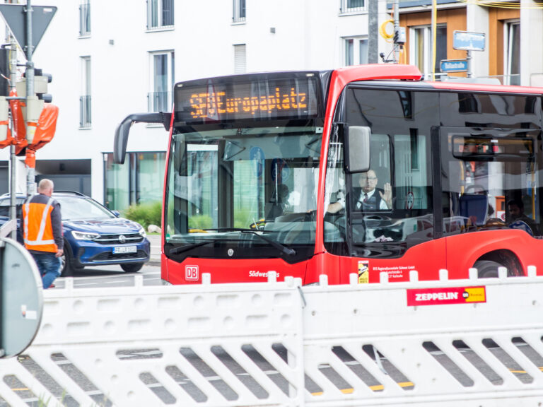 Red city bus with DB logo front and center, driver visible through the window, urban street scene with construction barriers nearby.