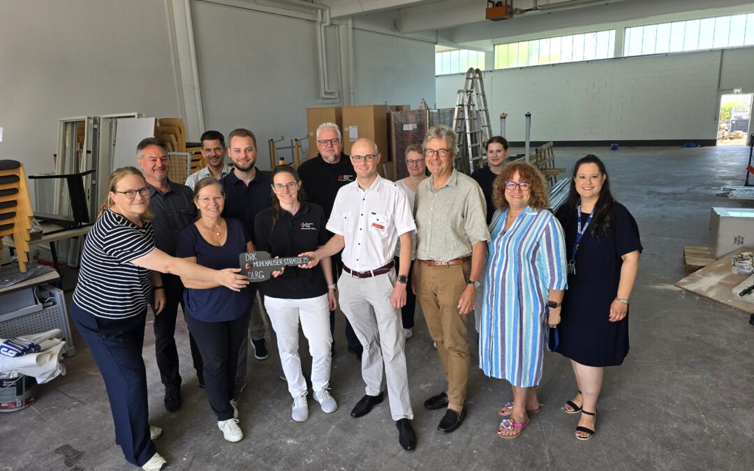 Group of adults posing together in a bright industrial workshop, with ladders, cabinets, and tools in the background.