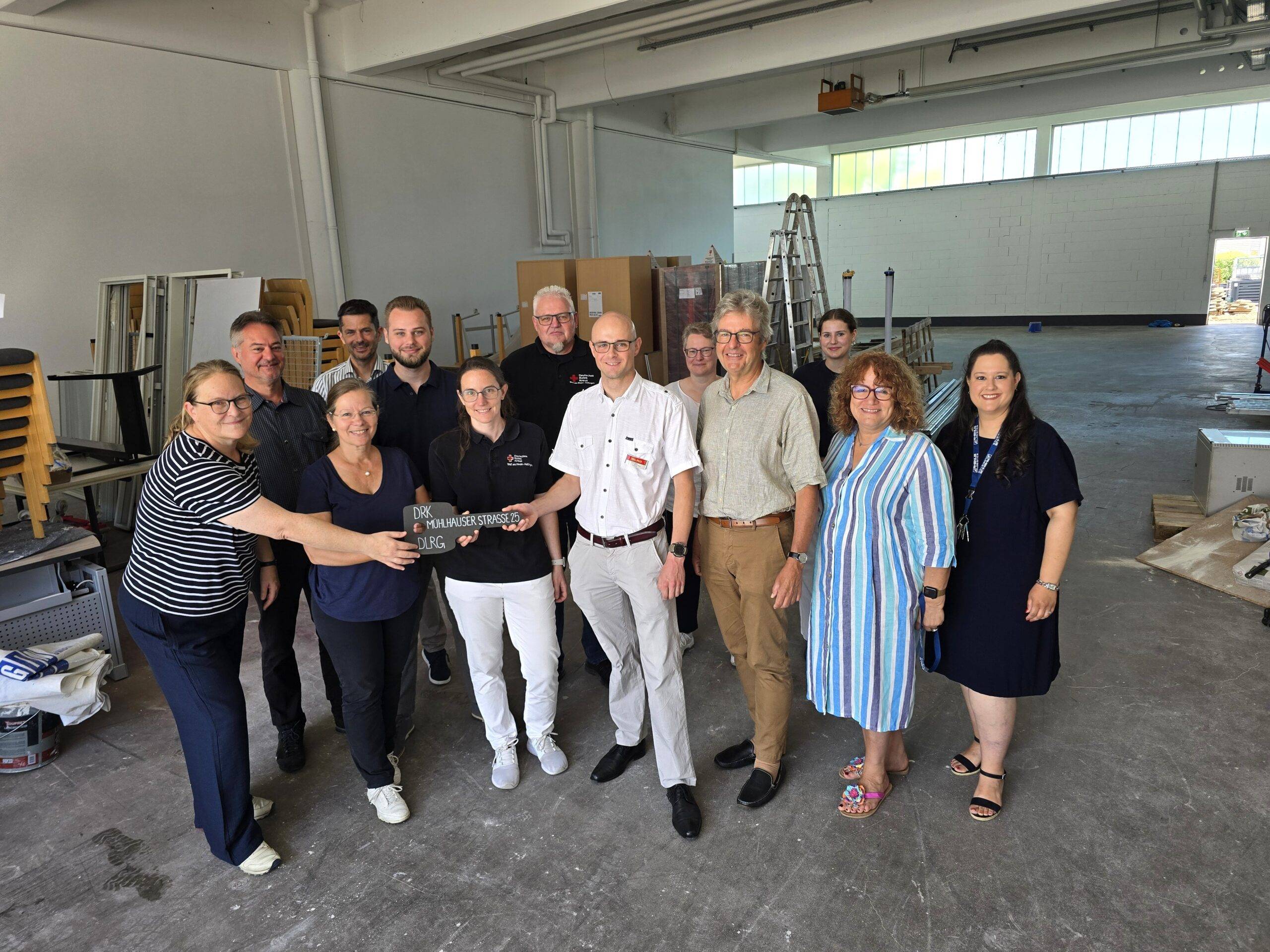 Group of adults posing together in a bright industrial workshop, with ladders, cabinets, and tools in the background.