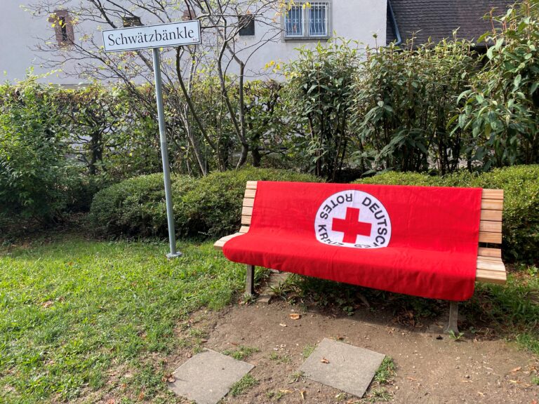 Red-covered wooden bench with a white circular logo featuring a red cross, placed in a garden with shrubs and a 'Schwätzbänkle' sign nearby.