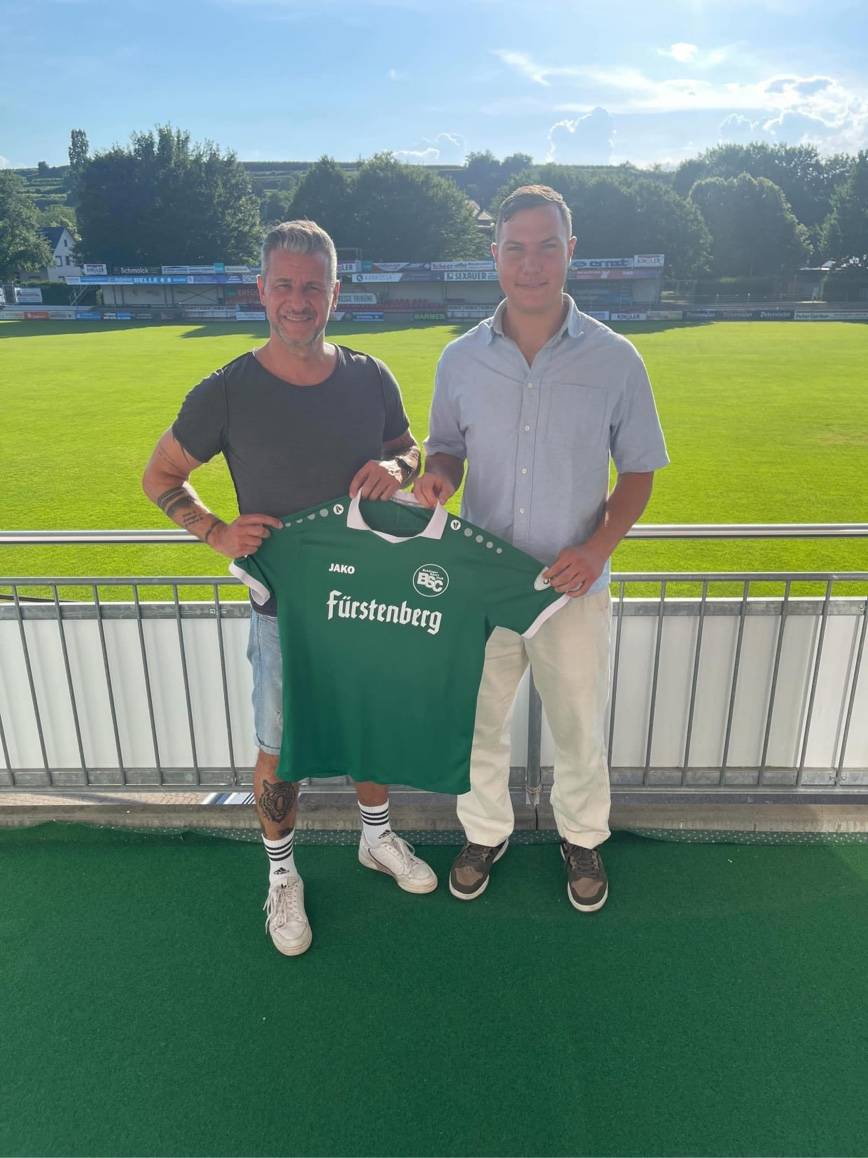 Two men pose on a stadium balcony, holding a green soccer jersey with 'Fürstenberg' logo, smiling with a sunny field behind them.