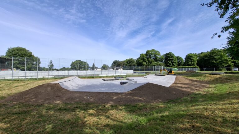 Wide view of an outdoor skatepark with concrete bowls, dirt ramps, and a chain-link fence, surrounded by trees under a blue sky.