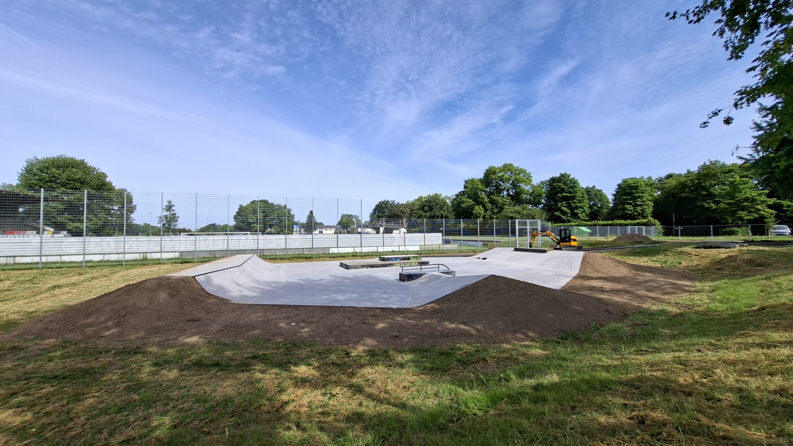 Wide view of an outdoor skatepark with concrete bowls, dirt ramps, and a chain-link fence, surrounded by trees under a blue sky.