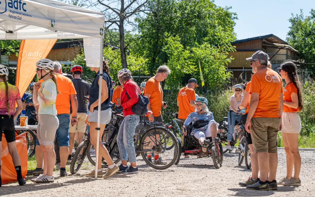 group of cyclists in orange shirts and helmets gather near a white adfc tent outdoors on a sunny day with bicycles nearby