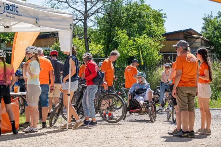 group of cyclists in orange shirts and helmets gather near a white adfc tent outdoors on a sunny day with bicycles nearby