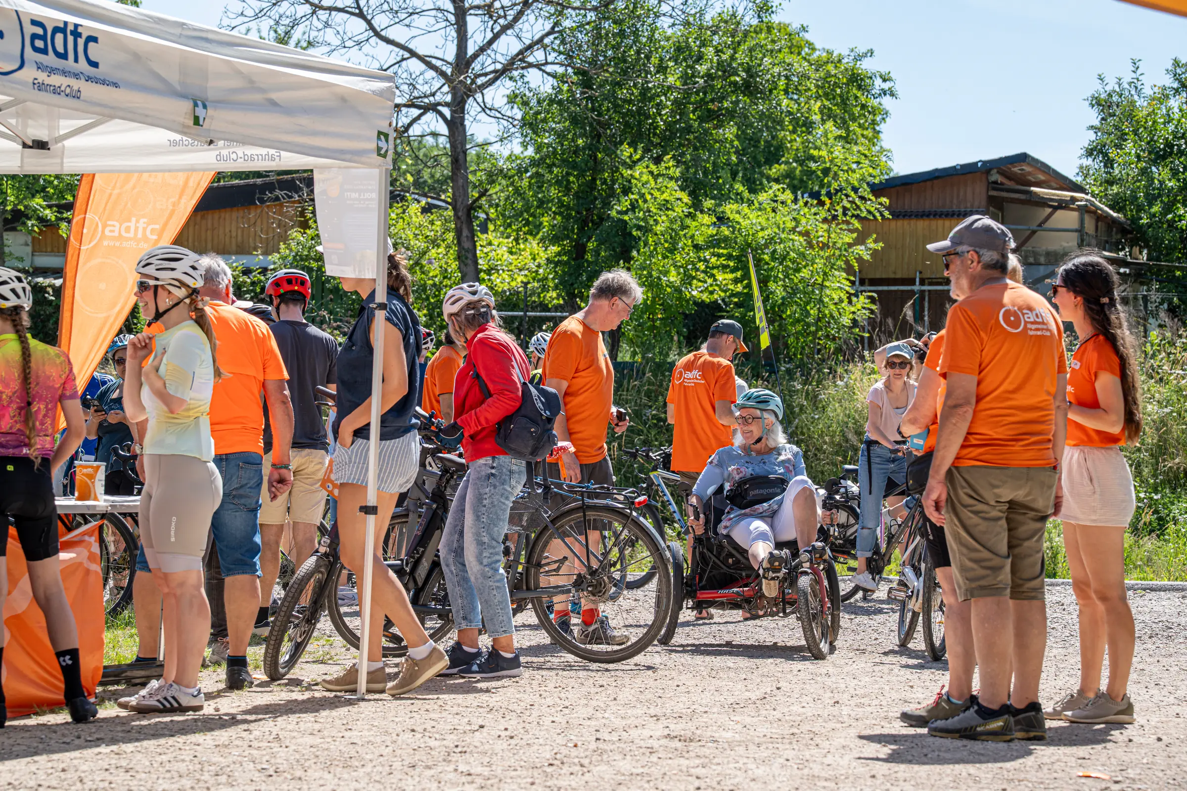 group of cyclists in orange shirts and helmets gather near a white adfc tent outdoors on a sunny day with bicycles nearby