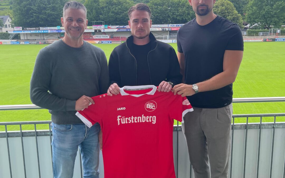Three men pose on a stadium balcony holding a red football jersey with 'Fürstenberg' text visible on the front.