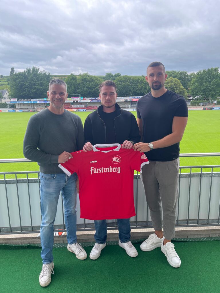 Three men pose on a stadium balcony holding a red football jersey with 'Fürstenberg' text visible on the front.