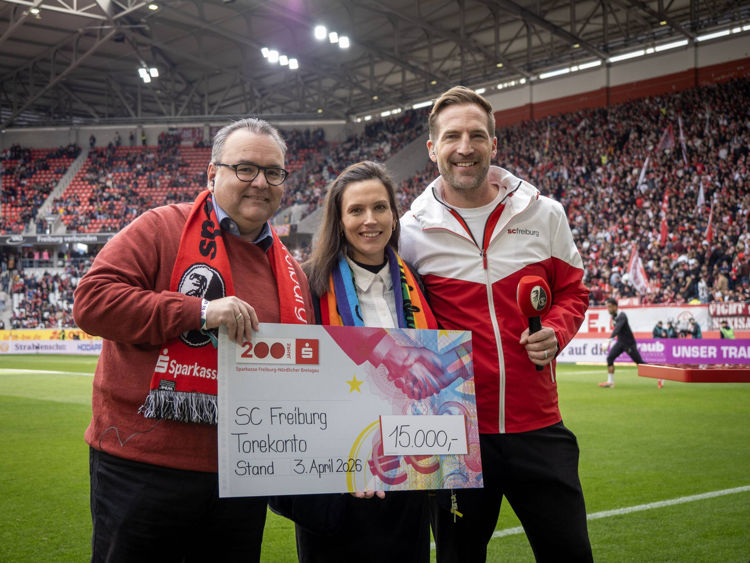 Three adults on a soccer field pose with a large colorful donation check, smiling, as a packed stadium watches from the stands behind them.