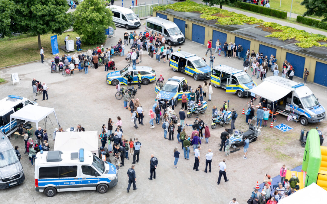 Aerial view of a public safety event with Polizei vans, a patrol car, tents, and a crowd of adults and children in a parking lot area.